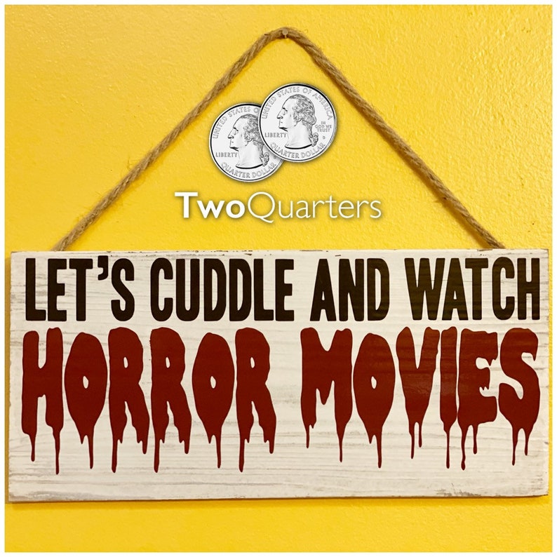 Op de afbeelding: Een wit houten bord met zwarte tekst die "Let's cuddle and watch horror movies" luidt. Het woord "horror" is in rode, druipende verf geschreven. Het bord hangt aan een touw met twee Amerikaanse kwartjes.