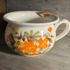 May include: A cream-colored ceramic chamber pot featuring a painted floral pattern with vibrant orange and yellow flowers, green leaves, and brown stems. The vessel has a wide, rounded body, a thick rim, and a single curved handle on the side.