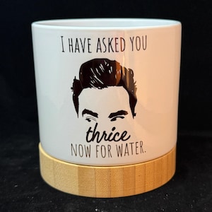 May include: White ceramic mug with a black graphic of a man's face and the text "I HAVE ASKED YOU thrice NOW FOR WATER." The mug sits on a round, light brown bamboo coaster. The mug is cylindrical.