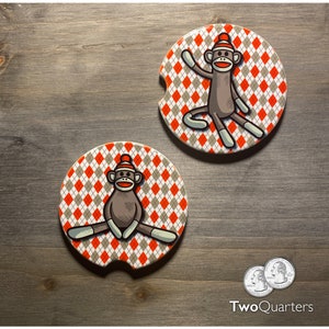 May include: Two round ceramic coasters with a red and white argyle pattern. Each coaster features a cartoon illustration of a monkey wearing a red and white stocking cap. The monkey on the left coaster is sitting with its legs crossed. The monkey on the right coaster is standing with one arm raised.