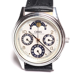 Może przedstawiać: Srebrny zegarek na rękę z białą tarczą, z wieloma tarczami pomocniczymi i wskaźnikiem faz księżyca. Zegarek ma czarne wskazówki i cyfry oraz czarny skórzany pasek. Widoczna jest nazwa marki "ORIS".