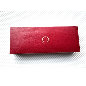 May include: A rectangular, red box with a textured surface. The box features a gold-colored Omega symbol in the center. The box is likely for a watch or other jewelry item.