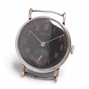 Op de afbeelding: Vintage polshorloge met een zwarte wijzerplaat en goudkleurige wijzers en cijfers. Het horloge heeft een kleine subwijzerplaat en een zilverkleurige metalen kast. Het woord "PRINCE" staat op de wijzerplaat. Het horloge is bevestigd aan een lichtgroene leren band.