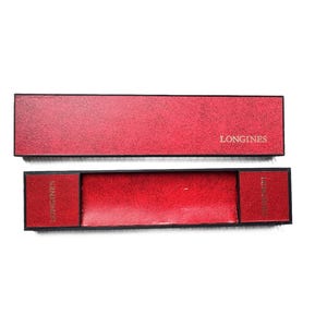 May include: Red rectangular watch box with the word "LONGINES" printed in gold. The box is open, revealing a red interior. The box is made of a textured material.