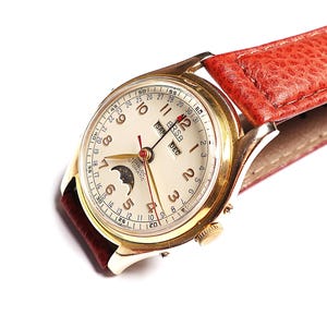 May include: A vintage gold-tone wristwatch with a cream-colored face and a red leather band. The watch face displays the time, a moon phase indicator, and the brand name. The band is textured and the watch is angled, showcasing its design.