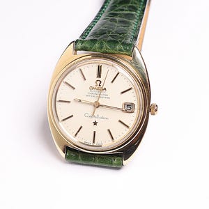 Könnte beinhalten: Eine goldfarbene Omega Constellation-Uhr mit cremefarbenem Zifferblatt, schwarzen Markierungen und Datumsfenster. Sie hat ein grünes Lederarmband. Auf dem Zifferblatt steht "Omega Automatic Chronometer Officially Certified Constellation".