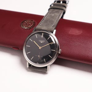 Könnte beinhalten: Eine silberfarbene Armbanduhr mit rundem schwarzem Zifferblatt, goldfarbenen Zeigern und einem grauen Lederarmband. Das Zifferblatt zeigt das Longines-Logo und ein kleines Hilfszifferblatt auf der Sechs-Uhr-Position. Sie liegt auf einem langen, glatten bordeauxroten Etui.