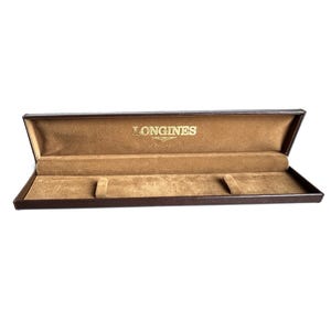 May include: An open, rectangular watch box with a brown exterior and a tan interior. The interior is lined with a soft material and features three cushioned compartments. The gold-colored brand name "LONGINES" is printed on the inside of the lid.