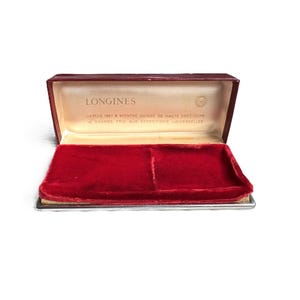 May include: An open, rectangular watch box with a burgundy exterior and a red velvet interior. The inside lid has the brand name "LONGINES" printed in gold lettering. The box is designed to hold a watch.
