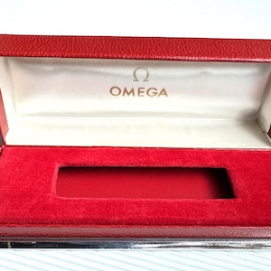 May include: Open red watch box with a silver base and a white interior. The inside is lined with red velvet and features the Omega logo in gold lettering. The box is designed to hold a watch.