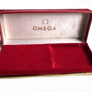 May include: Open red velvet watch box with gold trim. The interior features a cream-colored lining with the Omega logo in gold lettering. The box is designed to hold a watch.