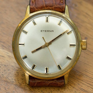 Puede incluir: Un reloj de pulsera Eterna dorado con esfera clara y agujas e indicadores dorados. El reloj tiene una correa de cuero marrón con un patrón de cocodrilo. El reloj se muestra sobre una superficie de madera.