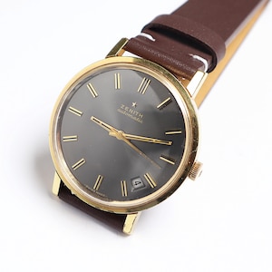 May include: A gold Zenith automatic wristwatch with a dark grey face and gold hands and markers. The watch has a brown leather strap. The watch face displays the date and the Zenith logo.