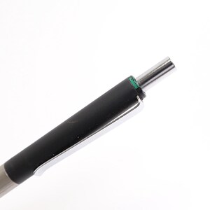 Puede incluir: Un bol&iacute;grafo plateado y negro con punta cromada y agarre negro. El bol&iacute;grafo tiene un peque&ntilde;o acento verde cerca de la punta. El bol&iacute;grafo se muestra sobre un fondo blanco.
