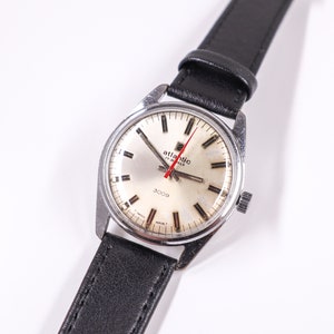 May include: A silver wristwatch with a white face and black leather band. The watch face has black markings and a red second hand. The watch face also has the text "Atlantic 17 Jewels 3009" printed on it.