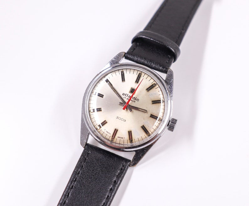May include: A silver wristwatch with a white face and black leather band. The watch face has black markings and a red second hand. The watch face also has the text "Atlantic 17 Jewels 3009" printed on it.
