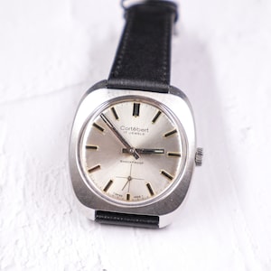 May include: A silver wristwatch with a white face and black leather band. The watch face has black markings and the text "Cortébert 17 Jewels Shockproof".