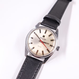 May include: A silver wristwatch with a white face and black leather band. The watch face has black Roman numeral hour markers, a red second hand, and the text "Atlantic 17 Jewels 3009" printed on it.
