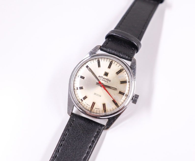 May include: A silver wristwatch with a white face and black leather band. The watch face has black Roman numeral hour markers, a red second hand, and the text "Atlantic 17 Jewels 3009" printed on it.