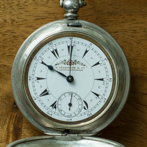 May include: A silver pocket watch with a white face and black hands. The watch face features black markers, a smaller subdial, and the text "K. SERKISOFF & CO CONSTANTINOPLE". The watch is open, revealing the inner workings.