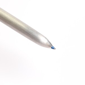 Puede incluir: Primer plano de un bol&iacute;grafo plateado con punta de tinta azul. El bol&iacute;grafo tiene un acabado met&aacute;lico y un dise&ntilde;o c&oacute;nico. La tinta azul es visible en la punta de escritura del bol&iacute;grafo. El bol&iacute;grafo est&aacute; sobre un fondo blanco.