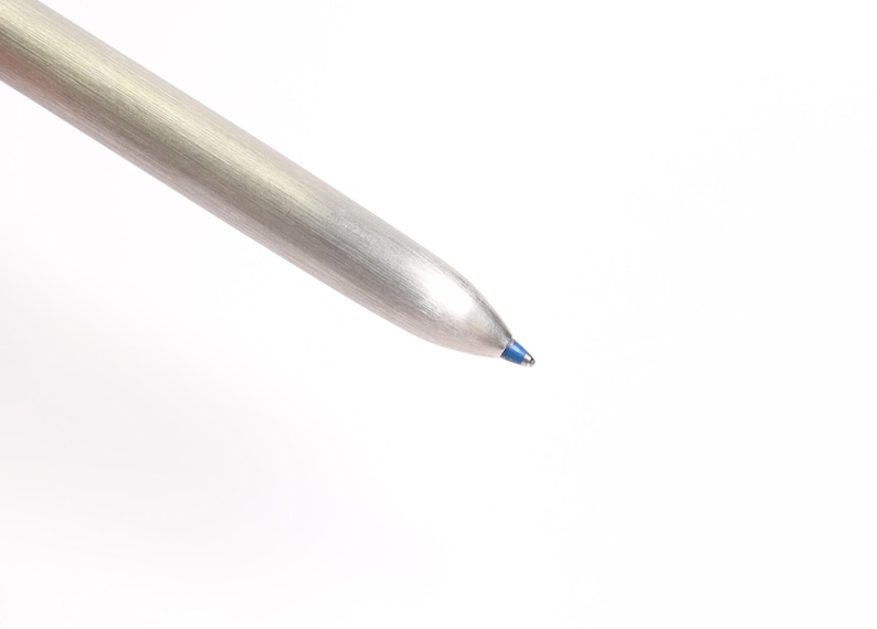 Puede incluir: Primer plano de un bol&iacute;grafo plateado con punta de tinta azul. El bol&iacute;grafo tiene un acabado met&aacute;lico y un dise&ntilde;o c&oacute;nico. La tinta azul es visible en la punta de escritura del bol&iacute;grafo. El bol&iacute;grafo est&aacute; sobre un fondo blanco.