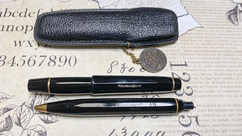 K&ouml;nnte beinhalten: Ein schwarzes Leder-Stift-Etui mit einer goldenen Kette und einem runden Anh&auml;nger mit den Olympischen Ringen und dem Text "XXII OLYMPIADE MUNCHEN 1972". Das Etui ist ge&ouml;ffnet und zwei schwarze Stifte sind sichtbar. Der Stift oben hat den Text "Hawecodport" an der Seite. Der Stift unten hat den Text "Hawecodport 619" an der Seite.