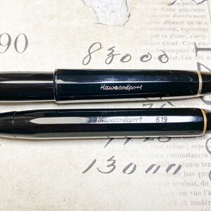 K&ouml;nnte beinhalten: Ein schwarzer Kaweco Sport F&uuml;llfederhalter und ein schwarzer Kaweco Sport Mechanik Bleistift. Beide Stifte haben eine goldene Verzierung und der Markenname "Kaweco Sport" ist in den Schaft graviert. Der mechanische Bleistift hat die Nummer "619" in den Schaft graviert.