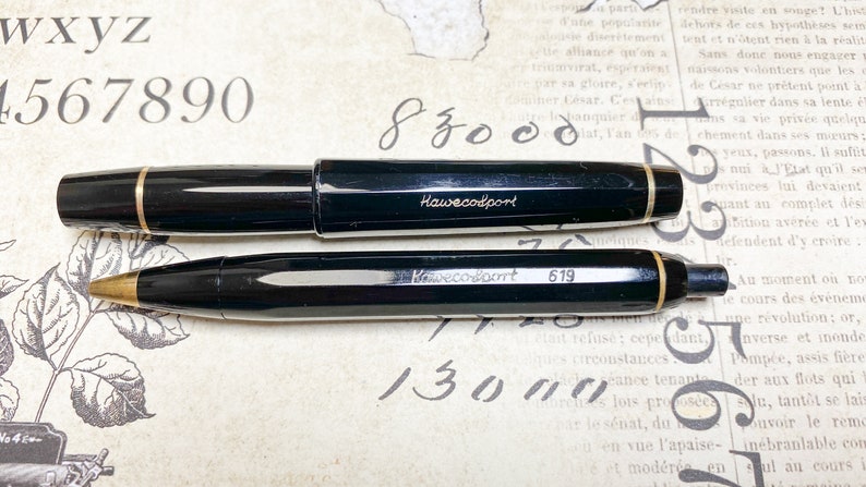 K&ouml;nnte beinhalten: Ein schwarzer Kaweco Sport F&uuml;llfederhalter und ein schwarzer Kaweco Sport Mechanik Bleistift. Beide Stifte haben eine goldene Verzierung und der Markenname "Kaweco Sport" ist in den Schaft graviert. Der mechanische Bleistift hat die Nummer "619" in den Schaft graviert.