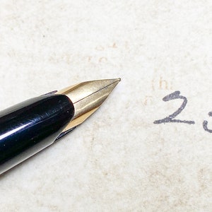 K&ouml;nnte beinhalten: Ein schwarzer und goldener F&uuml;llfederhalter mit einer feinen Feder, der auf einem Blatt Papier liegt, auf dem die Zahl "250" mit schwarzer Tinte geschrieben ist.