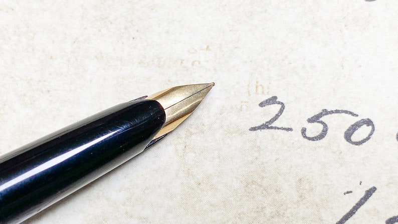 K&ouml;nnte beinhalten: Ein schwarzer und goldener F&uuml;llfederhalter mit einer feinen Feder, der auf einem Blatt Papier liegt, auf dem die Zahl "250" mit schwarzer Tinte geschrieben ist.