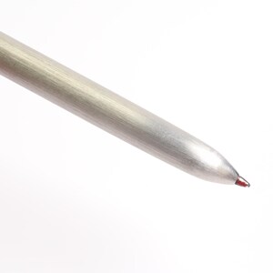 Puede incluir: Primer plano de un bol&iacute;grafo plateado con la parte superior negra. La punta del bol&iacute;grafo es de color rojo oscuro. El bol&iacute;grafo est&aacute; en &aacute;ngulo diagonal sobre un fondo blanco.