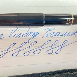 K&ouml;nnte beinhalten: Ein schwarzer und goldener F&uuml;llfederhalter mit der Aufschrift "V16 EF" auf der Kappe. Der Stift liegt auf einem Blatt liniertem Papier mit dem handschriftlichen Text "Nice Vintage Treasure" und einer Reihe von blauen Tintenklecksen.