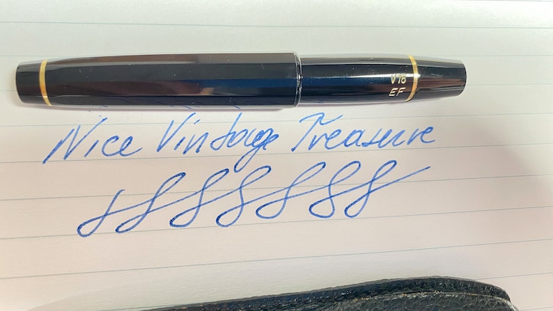 K&ouml;nnte beinhalten: Ein schwarzer und goldener F&uuml;llfederhalter mit der Aufschrift "V16 EF" auf der Kappe. Der Stift liegt auf einem Blatt liniertem Papier mit dem handschriftlichen Text "Nice Vintage Treasure" und einer Reihe von blauen Tintenklecksen.