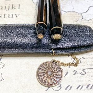 K&ouml;nnte beinhalten: Zwei schwarze Stifte mit goldenen Akzenten befinden sich in einem schwarzen Lederetui mit einer goldenen Kette und einem runden Anh&auml;nger. Der Anh&auml;nger hat ein Sonnenstrahlen-Design.