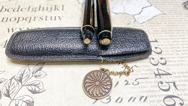 K&ouml;nnte beinhalten: Zwei schwarze Stifte mit goldenen Akzenten befinden sich in einem schwarzen Lederetui mit einer goldenen Kette und einem runden Anh&auml;nger. Der Anh&auml;nger hat ein Sonnenstrahlen-Design.