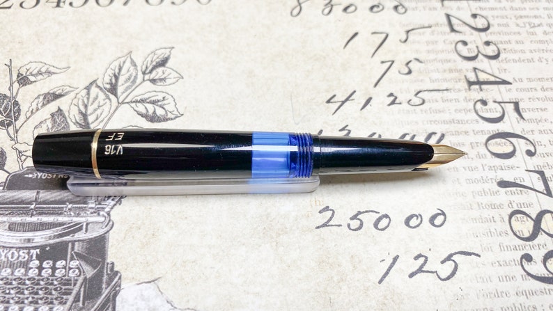 K&ouml;nnte beinhalten: Ein schwarzer und goldener F&uuml;llfederhalter mit einer blauen Tintenpatrone. Der F&uuml;ller hat eine goldene Verzierung und die Worte "V16 EF" sind auf der Seite eingraviert.