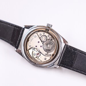 May include: A silver wristwatch with a black leather band. The back of the watch is open, revealing the inner workings of the watch. The watch face says "Swiss seventeen jewels".