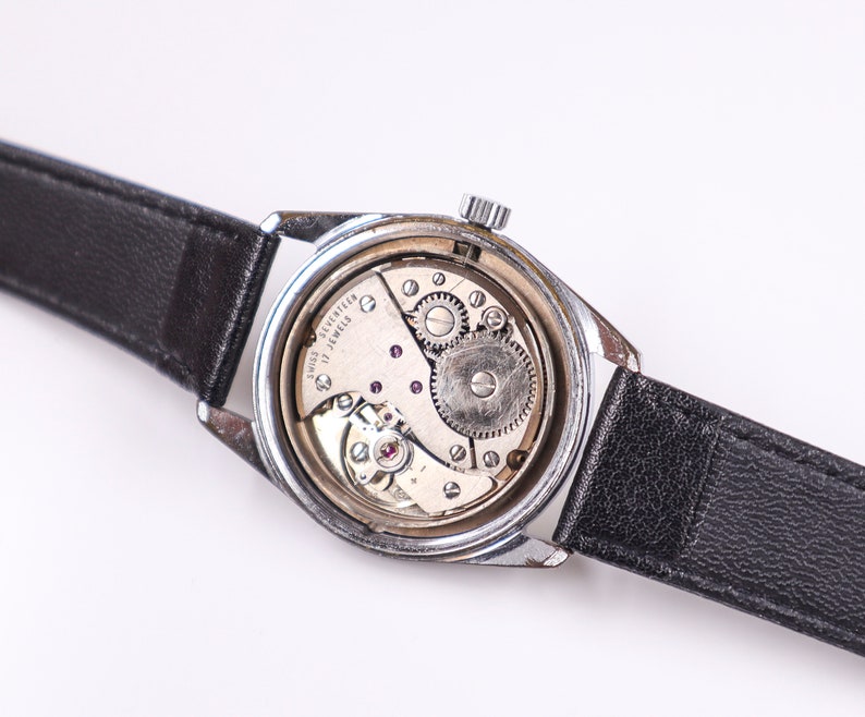 May include: A silver wristwatch with a black leather band. The back of the watch is open, revealing the inner workings of the watch. The watch face says "Swiss seventeen jewels".