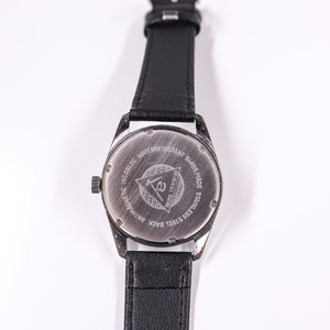 May include: The back of a silver watch with a black leather band. The watch face has the text "Incabloc Antimagnetic Waterresistant Swiss Made Stainless Steel Back Original Swiss" and a triangle with the letter "E" inside.