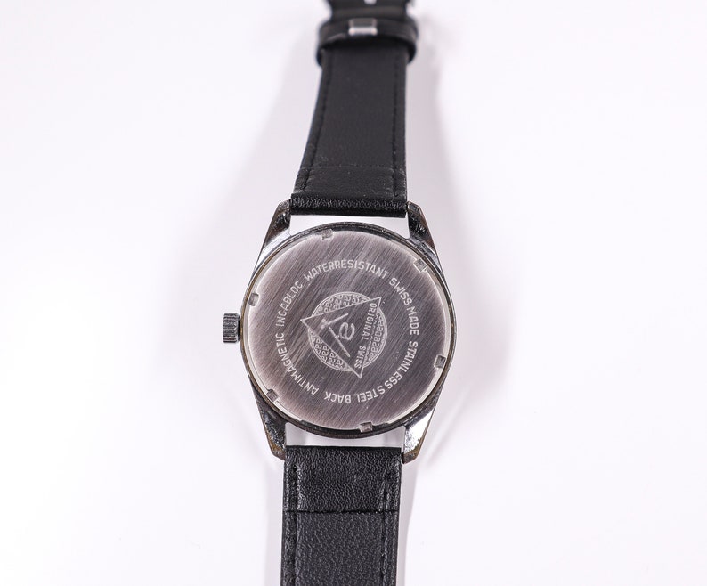 May include: The back of a silver watch with a black leather band. The watch face has the text "Incabloc Antimagnetic Waterresistant Swiss Made Stainless Steel Back Original Swiss" and a triangle with the letter "E" inside.