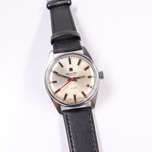 May include: A silver wristwatch with a white face and black leather band. The watch face has black markings and a red second hand. The watch face also has the text "Atlantic 17 Jewels 3009".