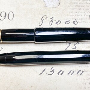 K&ouml;nnte beinhalten: Zwei schwarze Stifte mit goldenen Akzenten. Der gr&ouml;&szlig;ere Stift ist ein F&uuml;llfederhalter und der kleinere Stift ist ein Kugelschreiber. Die Stifte liegen auf einem Blatt Papier mit Schrift darauf.