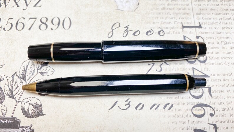 K&ouml;nnte beinhalten: Zwei schwarze Stifte mit goldenen Akzenten. Der gr&ouml;&szlig;ere Stift ist ein F&uuml;llfederhalter und der kleinere Stift ist ein Kugelschreiber. Die Stifte liegen auf einem Blatt Papier mit Schrift darauf.