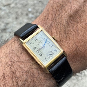 Könnte beinhalten: Vintage, goldfarbene rechteckige Armbanduhr mit cremefarbenem Zifferblatt und schwarzem Lederarmband. Das Zifferblatt der Uhr hat schwarze Ziffern, blaue Zeiger und den Text "Chronometre Titus". Die Uhr ist ca. 3,8 cm lang.