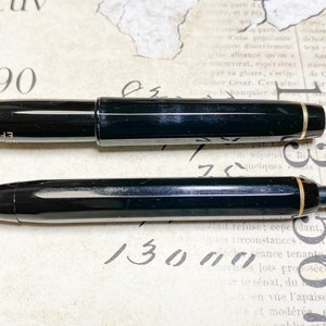 K&ouml;nnte beinhalten: Ein schwarzer F&uuml;llfederhalter und ein schwarzer Kugelschreiber mit goldenen Akzenten. Der F&uuml;llfederhalter hat eine schwarze Kappe und einen schwarzen Schaft mit einem goldenen Band. Der Kugelschreiber hat einen schwarzen Schaft mit einem goldenen Band und eine schwarze Kappe. Beide Stifte befinden sich auf einer wei&szlig;en Oberfl&auml;che.