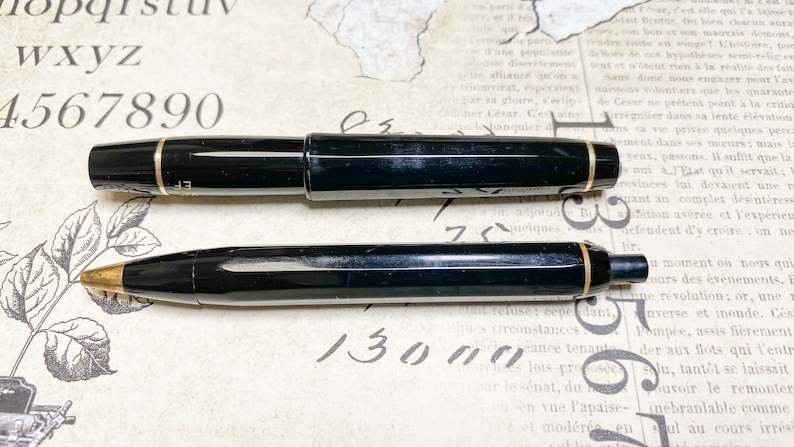 K&ouml;nnte beinhalten: Ein schwarzer F&uuml;llfederhalter und ein schwarzer Kugelschreiber mit goldenen Akzenten. Der F&uuml;llfederhalter hat eine schwarze Kappe und einen schwarzen Schaft mit einem goldenen Band. Der Kugelschreiber hat einen schwarzen Schaft mit einem goldenen Band und eine schwarze Kappe. Beide Stifte befinden sich auf einer wei&szlig;en Oberfl&auml;che.