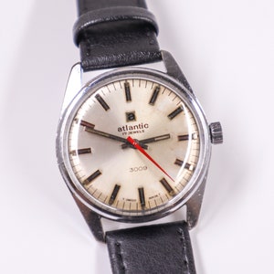 May include: A silver-toned wristwatch with a white face and black hands. The watch has a black leather band and says "atlantic 17 jewels 3009" on the face.