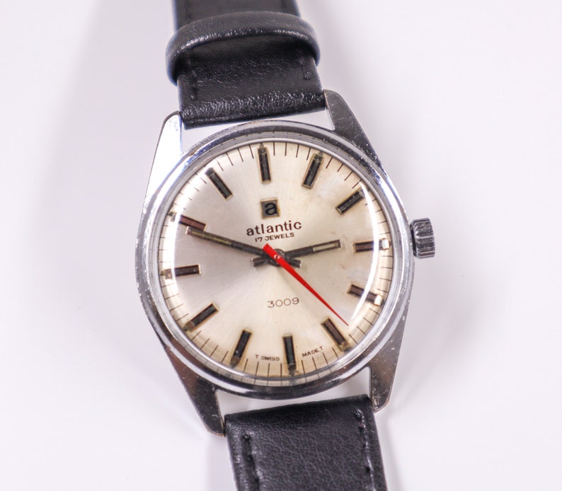 May include: A silver-toned wristwatch with a white face and black hands. The watch has a black leather band and says "atlantic 17 jewels 3009" on the face.
