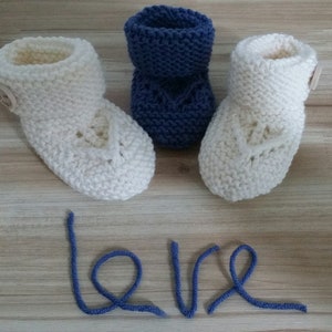 Puede incluir: Dos pares de botitas de bebé tejidas, una blanca con un botón y una azul. La palabra "love" está escrita en hilo azul sobre una superficie de madera.